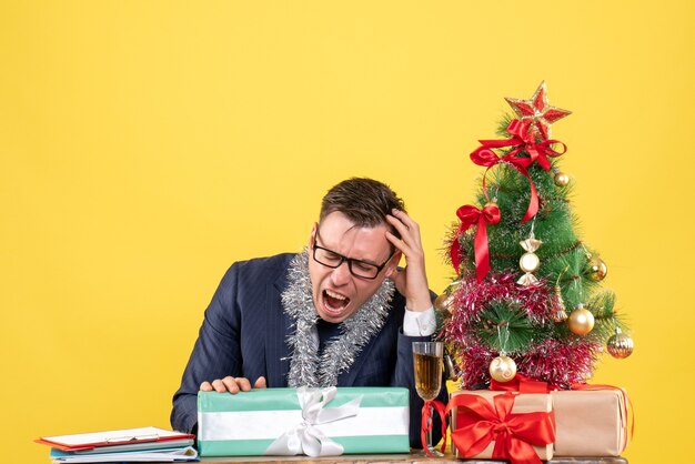 Vista frontal del hombre enojado sentado en la mesa cerca del árbol de Navidad y presenta sobre fondo amarillo