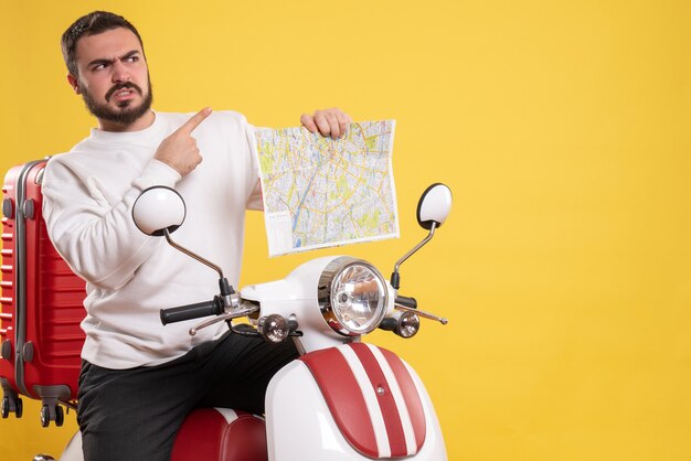 Vista frontal del hombre enojado sentado en la motocicleta con la maleta sosteniendo el mapa apuntando hacia arriba sobre fondo amarillo aislado