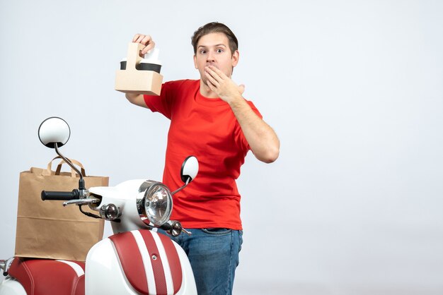 Vista frontal del hombre de entrega sorprendido en uniforme rojo de pie cerca de scooter mostrando orden sobre fondo blanco.