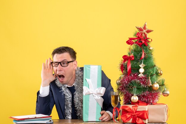 Vista frontal del hombre entrometido escuchando algo sentado en la mesa cerca del árbol de Navidad y presenta en amarillo