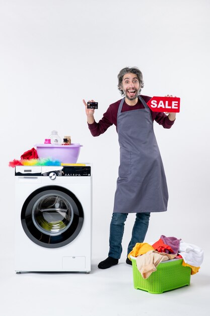 Vista frontal del hombre eufórico sosteniendo una tarjeta y un cartel de venta de pie cerca de la cesta de lavandería de la lavadora sobre fondo blanco.