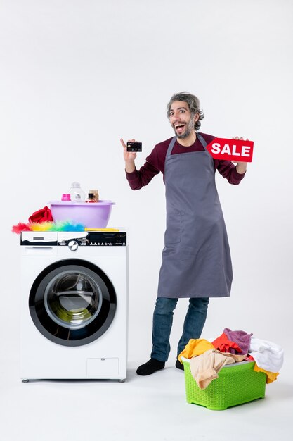 Foto gratuita vista frontal del hombre eufórico sosteniendo la tarjeta y el cartel de venta de pie cerca de la lavadora sobre fondo blanco.