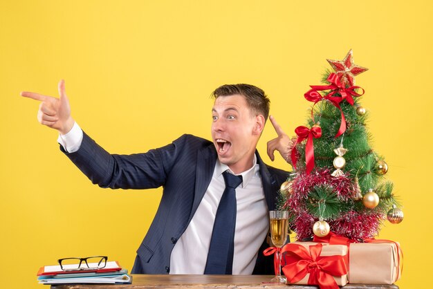 Vista frontal del hombre feliz apuntando con el dedo algo sentado en la mesa cerca del árbol de Navidad y presenta en amarillo