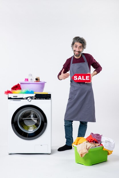 Vista frontal hombre feliz en delantal sosteniendo cartel de venta de pie cerca de la cesta de lavandería lavadora sobre fondo blanco.