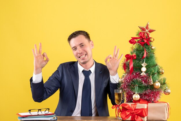 Vista frontal del hombre feliz haciendo letrero okey con las manos sentadas en la mesa cerca del árbol de Navidad y presenta en amarillo