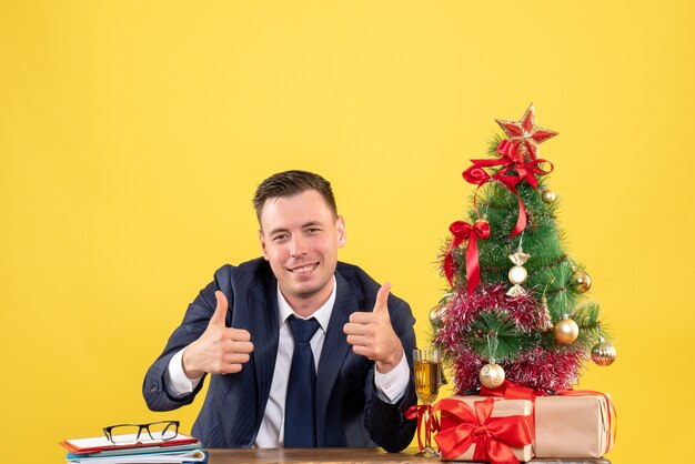 Vista frontal del hombre feliz haciendo el pulgar hacia arriba signo sentado en la mesa cerca del árbol de Navidad y regalos en amarillo