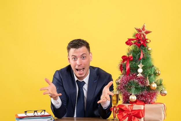 Vista frontal del hombre feliz con las manos abiertas sentado en la mesa cerca del árbol de Navidad y presenta en amarillo