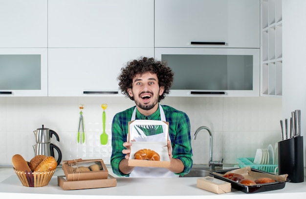 Vista frontal del hombre feliz mostrando pasteles recién horneados en una pequeña caja en la cocina blanca