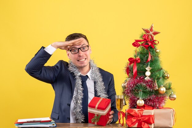 Vista frontal del hombre feliz poniendo la mano en la frente sentado en la mesa cerca del árbol de Navidad y presenta en amarillo