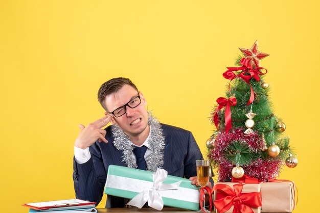 Vista frontal del hombre feliz poniendo la pistola de dedo en la sien sentado en la mesa cerca del árbol de Navidad y presenta en amarillo