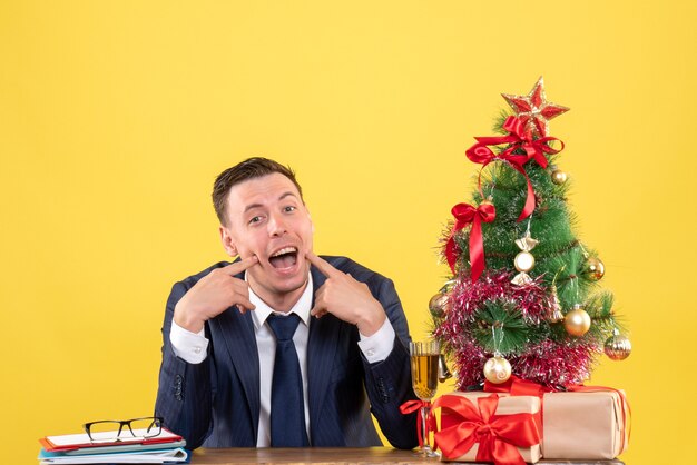 Vista frontal del hombre feliz sentado en la mesa cerca del árbol de Navidad y regalos en amarillo