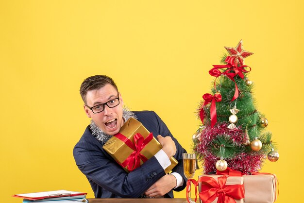 Vista frontal del hombre feliz sosteniendo su regalo firmemente en la mesa cerca del árbol de Navidad y presenta en amarillo