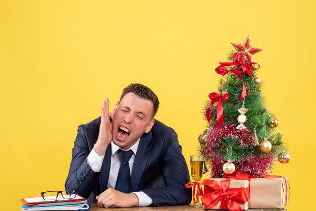 Vista frontal del hombre gritó sosteniendo su oreja sentado en la mesa cerca del árbol de Navidad y regalos en amarillo.