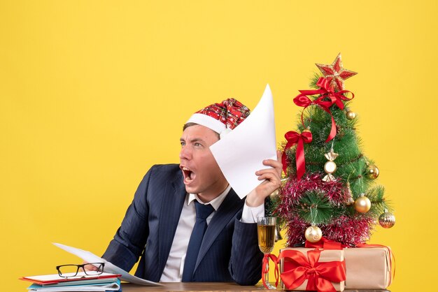 Vista frontal del hombre gritó tomando documentos sentado en la mesa cerca del árbol de Navidad y presenta en amarillo