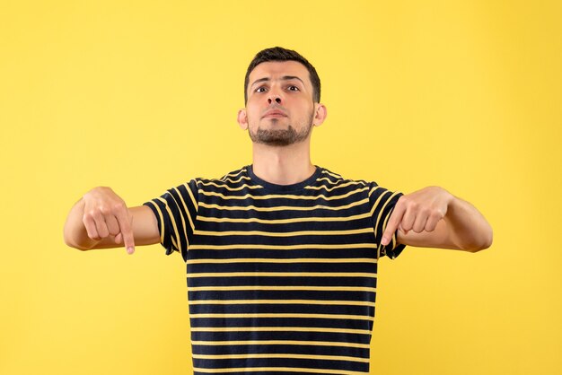 Vista frontal hombre guapo en camiseta a rayas blanco y negro apuntando al piso fondo amarillo aislado