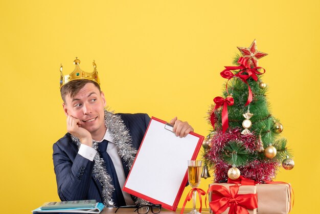 Vista frontal del hombre guapo con corona sentado en la mesa cerca del árbol de Navidad y presenta en amarillo