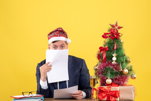 Vista frontal del hombre guapo con gorro de Papá Noel con documentos sentado en la mesa cerca del árbol de Navidad y regalos en amarillo