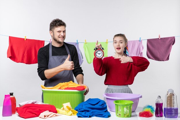 Vista frontal del hombre haciendo el pulgar hacia arriba y su esposa apuntando al reloj despertador de pie detrás de la mesa cestas de ropa y lavando cosas en la mesa