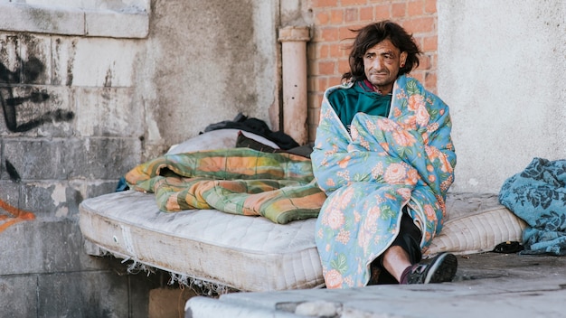 Vista frontal del hombre sin hogar en el colchón fuera debajo de una manta