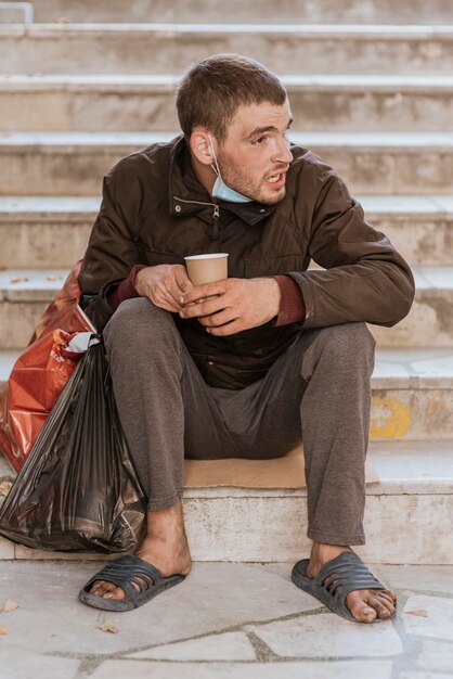 Vista frontal del hombre sin hogar en las escaleras con taza y bolsa de plástico