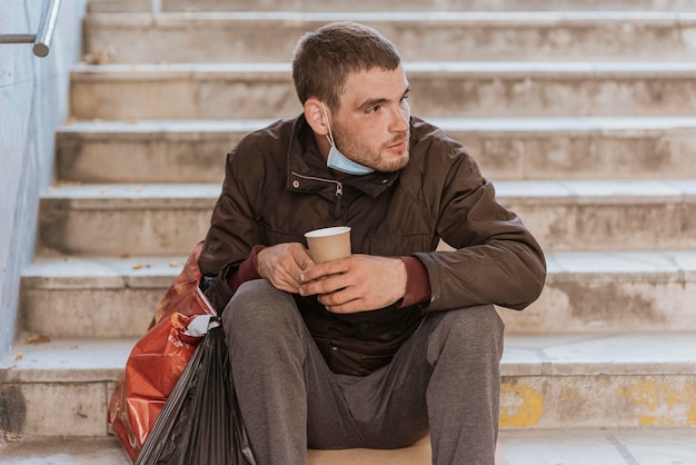 Vista frontal del hombre sin hogar sosteniendo una taza y una bolsa de plástico en las escaleras