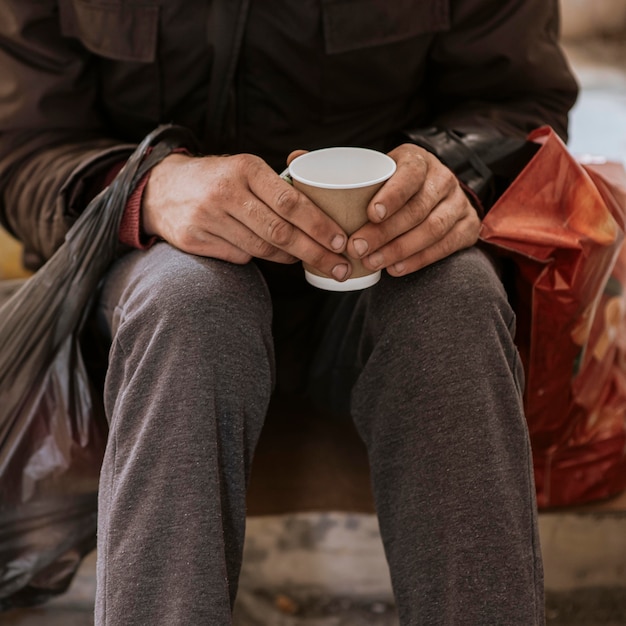 Vista frontal del hombre sin hogar sosteniendo una taza y una bolsa de plástico