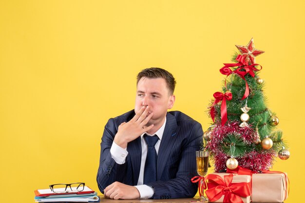 Vista frontal del hombre insatisfecho sentado en la mesa cerca del árbol de Navidad y presenta en amarillo