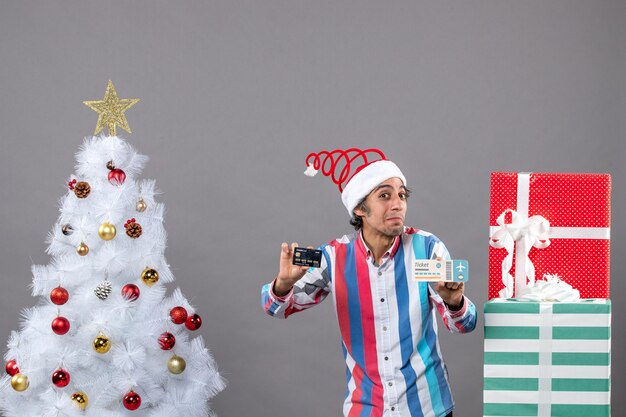 Vista frontal hombre interesado con tarjeta y boleto de viaje alrededor del árbol de Navidad y regalos