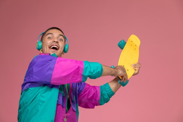 Vista frontal del hombre joven en abrigo colorido sosteniendo patineta sobre fondo rosa