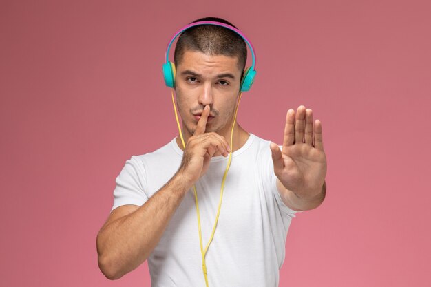 Vista frontal del hombre joven en camiseta blanca escuchando música showign signo de silencio en el escritorio rosa