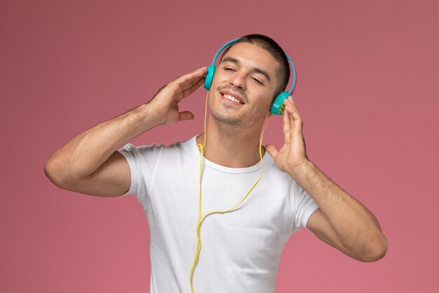 Vista frontal del hombre joven en camiseta blanca escuchando música a través de auriculares en el escritorio rosa