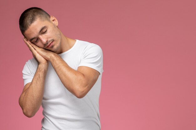 Vista frontal del hombre joven en camiseta blanca posando en expresión de dormir sobre fondo rosa