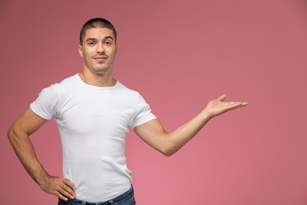 Vista frontal del hombre joven en camiseta blanca posando con la mano levantada y la palma sobre fondo rosa