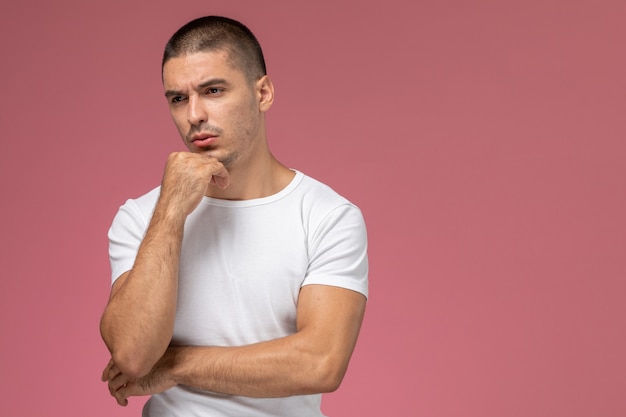 Vista frontal del hombre joven en camiseta blanca posando y pensando sobre fondo rosa