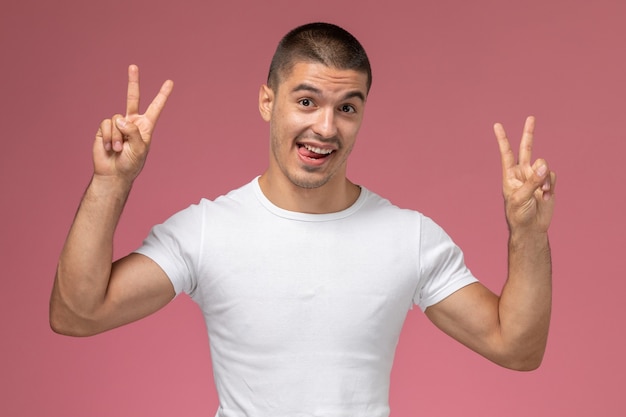 Vista frontal del hombre joven en camiseta blanca posando con signos de victoria sobre fondo rosa