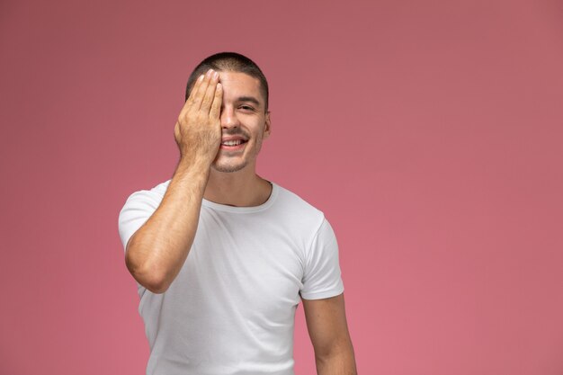 Vista frontal del hombre joven en camiseta blanca que cubre la mitad de su rostro con una sonrisa en el escritorio rosa