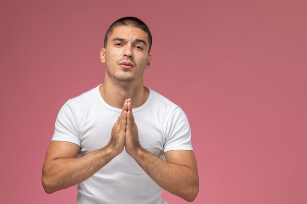 Vista frontal del hombre joven en camiseta blanca rezando posando sobre fondo rosa