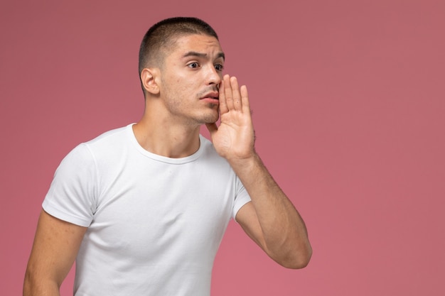 Vista frontal del hombre joven en camiseta blanca susurrando sobre fondo rosa