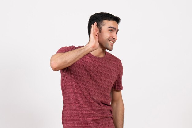 Vista frontal del hombre joven en camiseta de color rojo oscuro tratando de escuchar sobre fondo blanco.