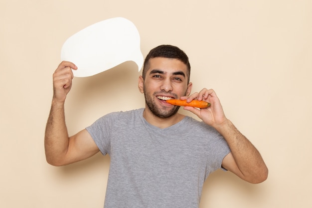 Vista frontal del hombre joven en camiseta gris con cartel blanco y comer zanahoria en beige