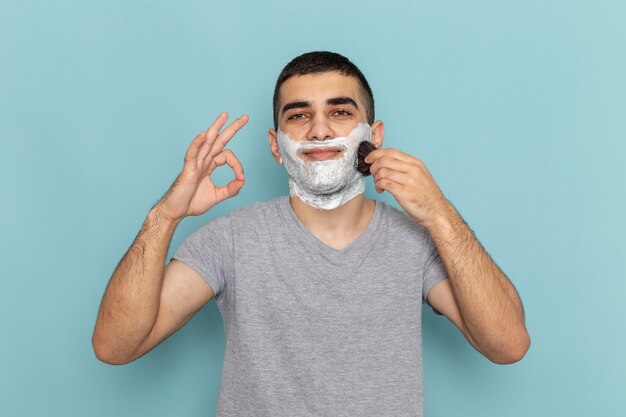 Vista frontal del hombre joven en camiseta gris con espuma blanca en la cara frotándola con un pincel sobre azul hielo