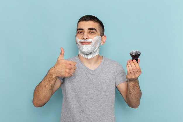 Vista frontal del hombre joven en camiseta gris con espuma blanca en la cara sosteniendo el cepillo en azul helado