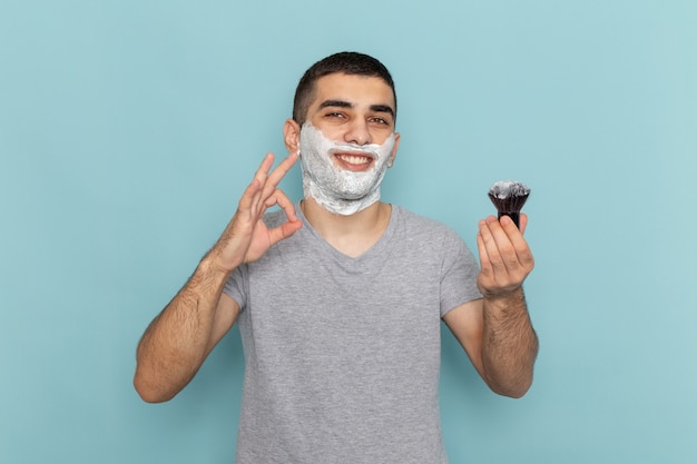 Vista frontal de un hombre joven en camiseta gris con espuma blanca en su rostro sonriendo en azul hielo