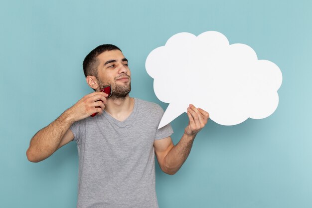 Vista frontal hombre joven en camiseta gris con un gran cartel blanco y maquinilla de afeitar eléctrica en la pared azul hielo barba espuma afeitadora de pelo