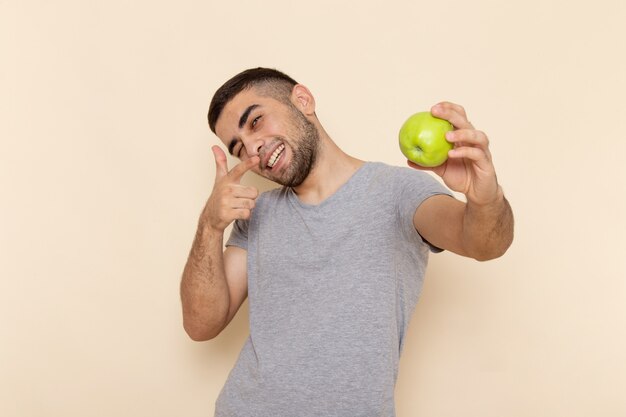 Vista frontal del hombre joven en camiseta gris y jeans azules sonriendo y sosteniendo manzana verde en beige