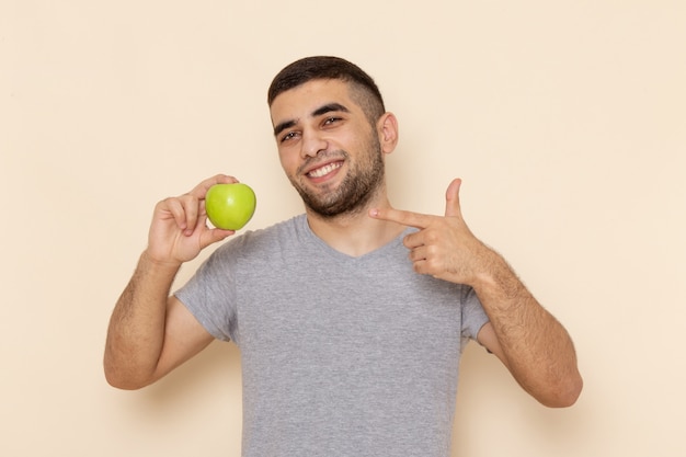 Vista frontal del hombre joven en camiseta gris y jeans azules sonriendo y sosteniendo manzana verde en beige
