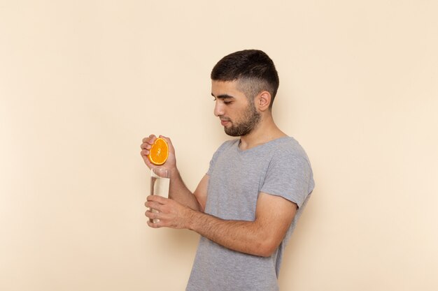 Vista frontal hombre joven en camiseta gris y jeans exprimiendo jugo de naranja en beige