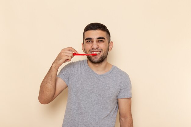 Vista frontal hombre joven en camiseta gris limpiando sus dientes en beige