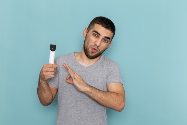 Vista frontal hombre joven en camiseta gris con maquinilla de afeitar eléctrica en el escritorio azul barba de afeitar espuma de pelo masculino