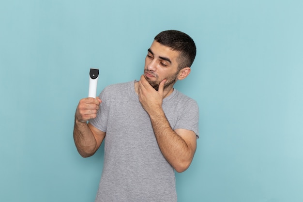 Vista frontal del hombre joven en camiseta gris con maquinilla de afeitar eléctrica pensando en la espuma de pelo masculino de barba azul afeitado